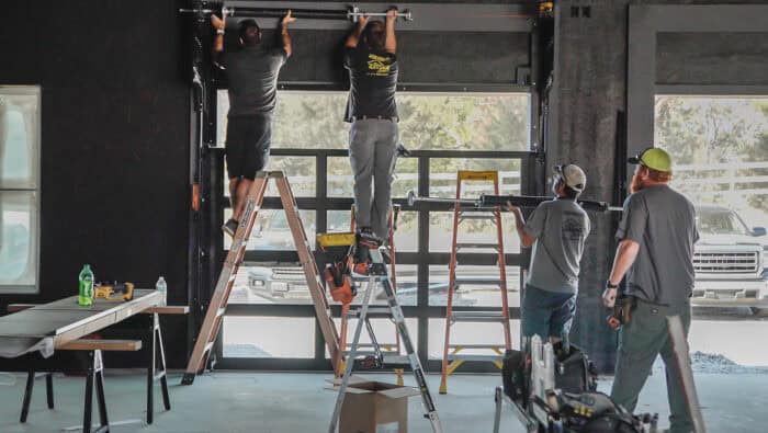 Four men install a garage door using ladders and tools. A truck is visible outside.