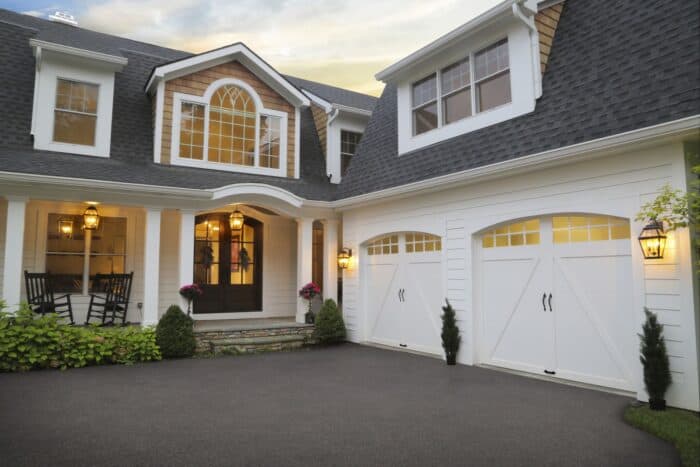 Front view of a large house with a double garage, white siding, and black trim. Two lanterns illuminate the entrance, and a rocking chair and potted plants decorate the porch.