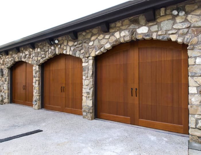 A stone wall showcases three wooden garage doors with black handles and arched tops, highlighting expert garage door installation.