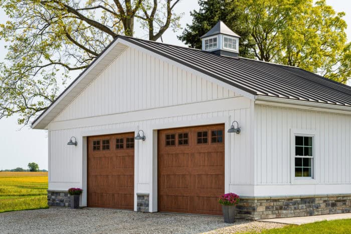 A white garage with two wooden doors, a gravel driveway, and potted plants in front. Trees and a field are in the background.