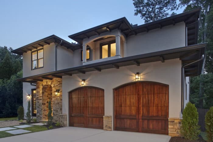A two-story house with a modern design, featuring stone accents and wooden double garage doors. It is surrounded by greenery and illuminated by exterior lights.