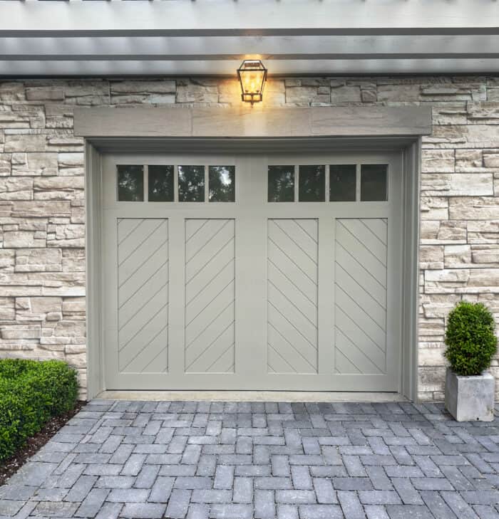 A residential garage door with diagonal panels and small windows at the top is set in a stone wall, complemented by a lantern above and potted plants on either side.