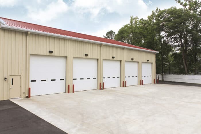 A beige warehouse with four residential garage doors, each framed by red bollards, sits on a spacious concrete lot. Trees and a white fence enhance the backdrop.